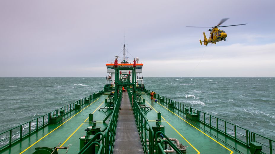 Ett grönt bulkfartyg, bild tagen från bryggan ut över däck med havet i bakgrunden. En gul helikopter syns i luften till höger om fartyget. 