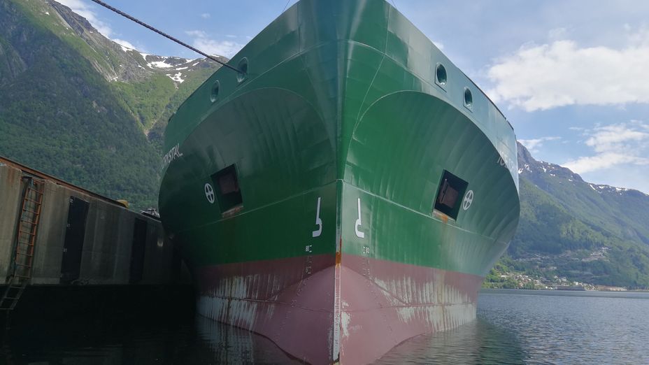Ett grönt bulkfartyg med rött skrov där bilden är tagen mot fören sett underifrån, med berg i bakgrunden.