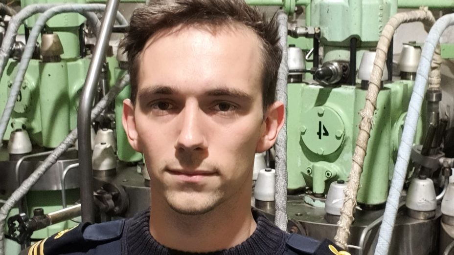 En porträttbild av förste fartygingenjören Martin i uniform i maskinrummet framför en av motorerna.