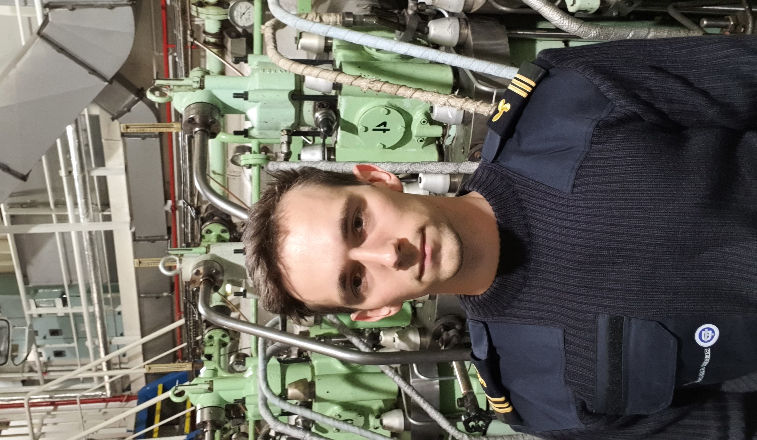 En porträttbild av förste fartygingenjören Martin i uniform i maskinrummet framför en av motorerna.